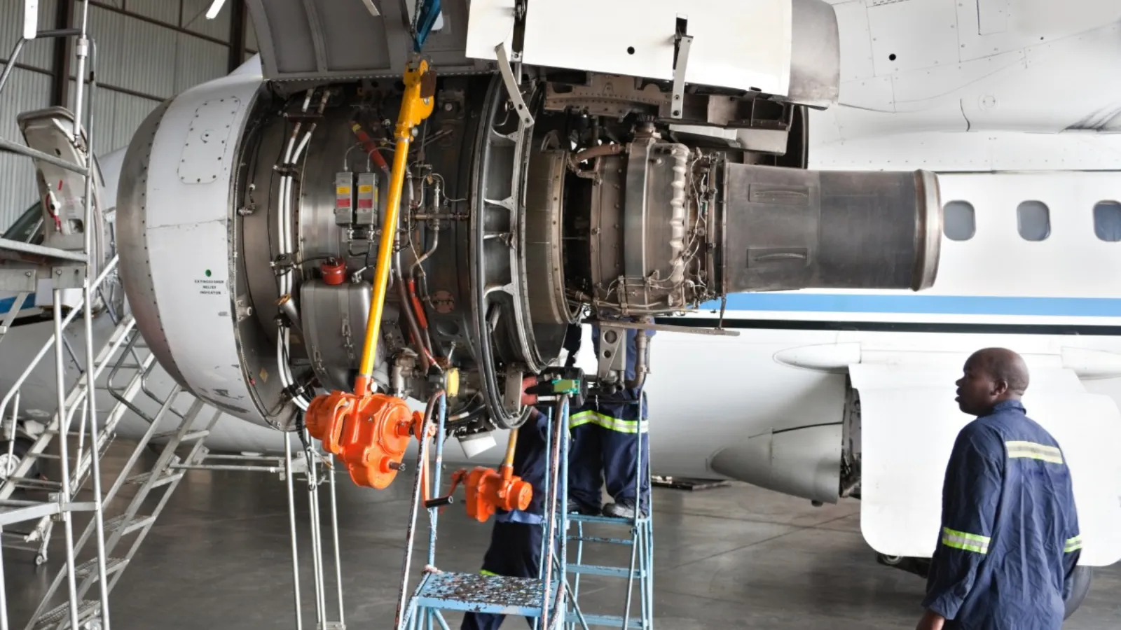 An aircraft engine opened up for maintenance in a hangar, with engineers in overalls working beneath it on step platforms.